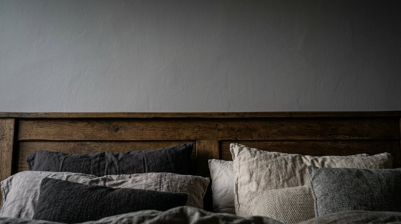 Close up of a wooden bed frame with stacked grey and beige linen pillows against a solid grey wall.